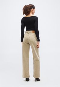 Γυναίκα με σγουρά μαλλιά που φοράει μαύρη μπλούζα με μακριά μανίκια, μπεζ τζιν με φαρδιά πόδια και μαύρες μπότες με ψηλό τακούνι, στέκεται με την πλάτη προς τα εμάς.