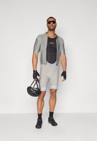 Tenue de cyclisme avec un maillot gris à manches courtes, un sous-vêtement en mesh noir, un short gris, des gants sans doigts et un casque noir tenu à la main.