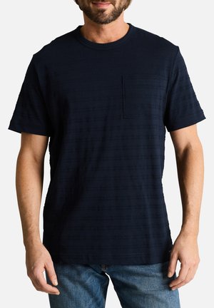 Homme portant un t-shirt bleu marine foncé à manches courtes avec une poche poitrine et un jean bleu, debout devant un fond clair uni.