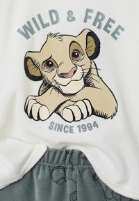 White t-shirt featuring an illustrated lion cub with large eyes, text "WILD & FREE" in gray, paired with gray shorts with a pattern.