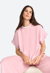 Pink oversized t-shirt with short sleeves and a round neck, made of smooth fabric. Worn with matching pink pants, seated on a light surface.