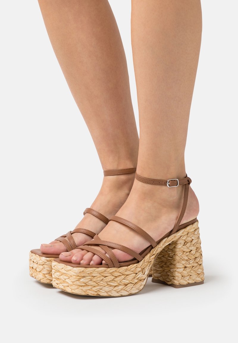 Pieds portant des sandales à plateforme marron à brides, avec talons texturés tissés et brides à boucle à la cheville sur fond blanc.