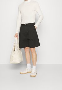 Pantalones cortos negros con pliegues, blusa de manga larga beige, zapatillas blancas con suelas marrones y un bolso de mano color crema. Diseño minimalista y colores neutros.
