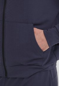 Marinblå zip-hoodie i mjukt tyg, med en framficka, ribbade muddar och en slät yta. Dekorativ sömnad syns.