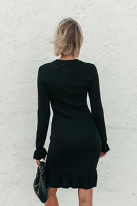 Schwarzes Strickkleid mit langen Ärmeln, geripptem Ausschnitt und gerüschtem Saum. Das Modell hält eine gewebte schwarze Handtasche. Strukturiertes Stoffdetail sichtbar.