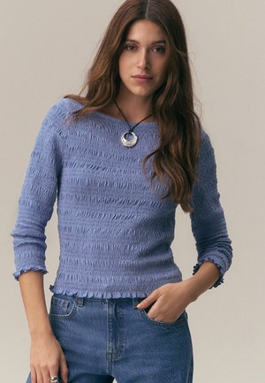 Young woman with long brown hair wears a textured light blue long-sleeve top, blue jeans, and a round silver pendant necklace.