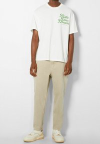 T-shirt bianca in cotone con testo verde "Rendi i tuoi sogni" sul petto, abbinata a pantaloni beige e sneakers bianche con accenti chiari.