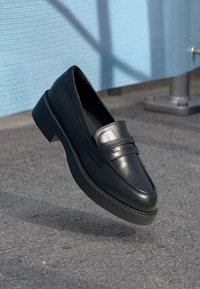 Mocassins en cuir noir avec une finition lisse, présentant un détail de bande en relief sur le dessus et une semelle épaisse et texturée. Design élégant.