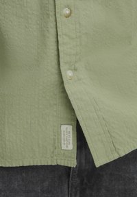 Camicia button-up verde chiaro con tessuto texture, bottoni in perla e etichetta tessuta sull'orlo, abbinata a pantaloni grigio scuro.