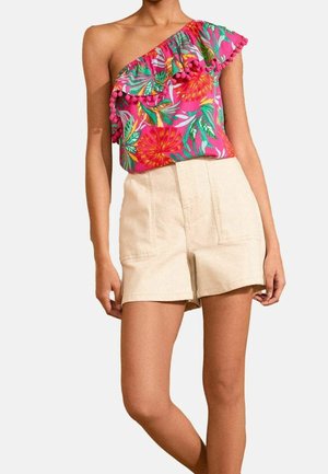 Femme portant un haut rose à fleurs à une épaule avec volants et pompons, associé à un short beige taille haute avec poches avant.