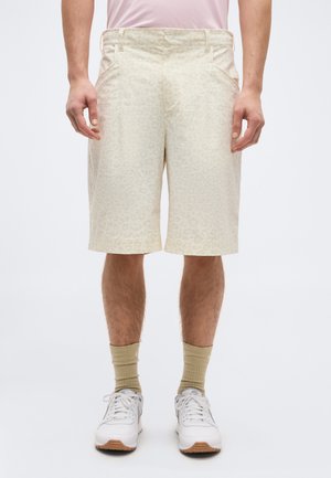 Homme portant un short beige à motifs jusqu'aux genoux, des chaussettes beiges, des baskets blanches et une chemise rose clair, debout devant un fond uni.