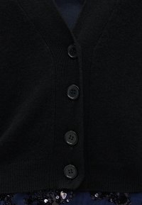 Cardigã de malha preta com decote em V, cinco botões redondos pretos e uma textura suave. O fundo inclui lantejoulas escuras.