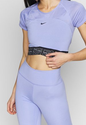 Femme portant un crop top sportif Nike Pro violet clair et des leggings taille haute assortis, tenant le bas du crop top.