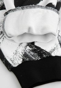 Czarny i biały detal bluzy z teksturowanym białym obszarem na ramieniu, czarnym ściągaczem oraz abstrakcyjnymi wzorami przypominającymi farbę na tkaninie.