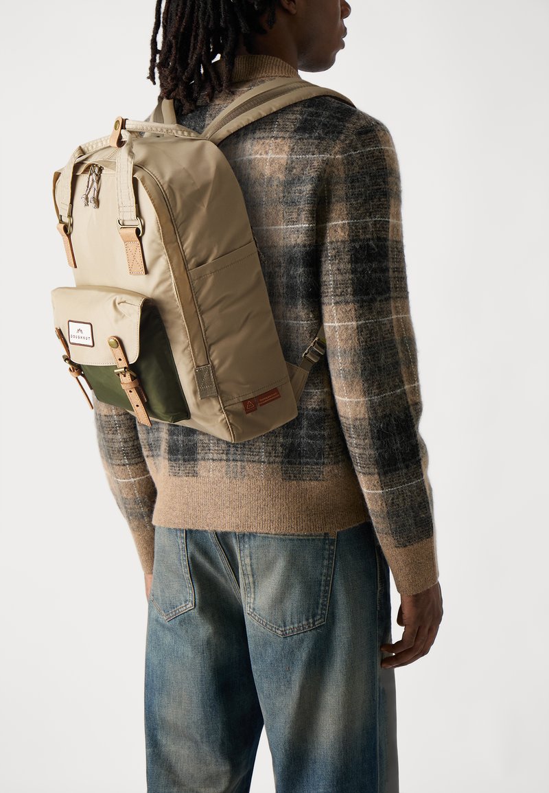 Tan backpack with green accents, featuring a top handle, adjustable straps, and brown leather details. Worn over a patterned sweater.