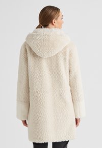 Frau in einem strukturierten cremefarbenen Kapuzenmantel mit hochgeschlagenen Ärmeln, von hinten vor einem schlichten Hintergrund gezeigt.