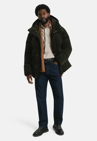 Gepolsterte dunkelgrüne Jacke mit Kapuze, beigefarbene Knopfleiste-Bluse, karierter Schal, dunkle Jeans und schwarze Schnürschuhe.