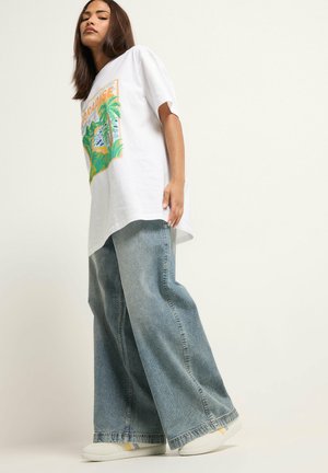 Mujer con camiseta blanca gráfica de gran tamaño, vaqueros anchos azules y zapatillas blancas, de pie frente a un fondo liso.