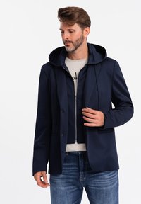 Marineblå to-i-én jakke, der kombinerer blazerstil med en hætte indvendig. Har glat stof, lynlås lukning og en afslappet pasform.