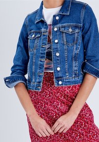 Veste en denim à manches longues, col classique, poches avant et boutons argentés. Portée sur un tee-shirt graphique et une jupe rouge à fleurs.