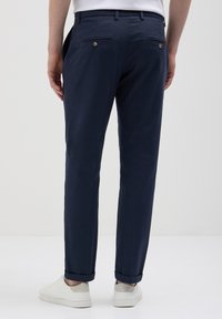 Uomo che indossa pantaloni slim-fit blu navy e sneakers bianche, in piedi contro uno sfondo chiaro e semplice, visto da dietro.