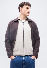 Νεαρός άνδρας φορώντας ένα σκούρο καφέ μπουφάν με φερμουάρ πάνω από ένα ελαφρύ μπεζ πλεκτό με φερμουάρ και μπλε τζιν, στέκεται με τα χέρια στις τσέπες, κοιτάζοντας δεξιά.