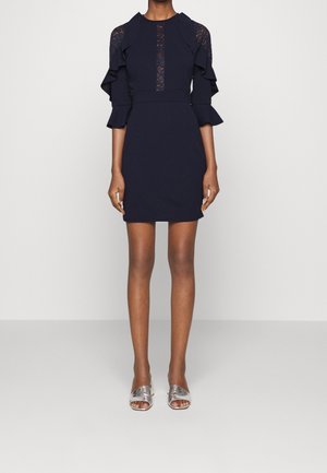 Kvinde iført en marineblå kjole med blonderpaneler og flæseærmer i halv længde, kombineret med sølvfarvede åbne slip-on sko.