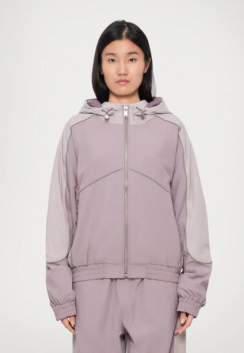 Ανοιχτό μωβ φούτερ με κουκούλα και φερμουάρ μπροστά, ελα�στικούς καρπούς και ελαφρώς χαλαρή εφαρμογή. Περιέχει αντίθετους γκρι τόνους και ορατές ραφές.