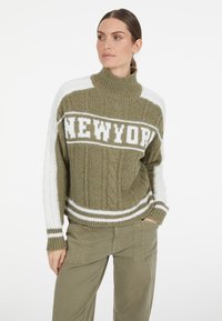 Olivgrüner, gestrickter Pullover mit hohem Kragen, der ein weißes "NEWYORK"-Grafikdesign, weiße Streifen und eine Zopfstricktextur an den Ärmeln aufweist.