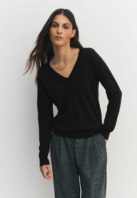 Pulover negru cu decolteu în V, realizat dintr-un material moale, cu mâneci lungi, croială lejeră și tiv reiat, asortat cu pantaloni gri cu model.