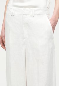 Personne portant un pantalon en lin blanc taille haute avec des poches latérales, une main reposant dans une poche, sur un fond uni.