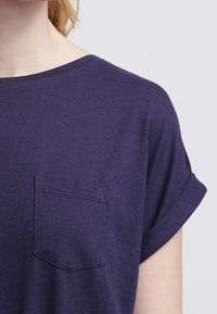 Fioletowa koszulka z krótkim rękawem, wykonana z miękkiego materiału, z przednią kieszonką i podwiniętymi rękawami, o luźnym dekolcie i gładkiej fakturze.