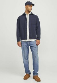 Chaqueta de algodón azul marino sobre una camisa blanca a rayas, combinada con jeans de color azul claro y zapatos de ante marrones. La chaqueta cuenta con un bolsillo en el pecho.