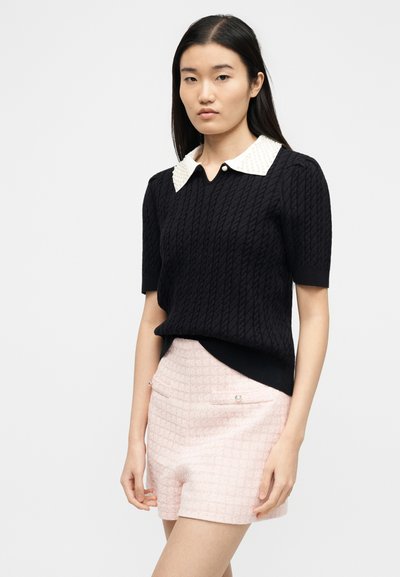 Femme portant un haut noir en maille à manches courtes avec un col orné de perles blanches et un short rose clair texturé avec des poches décoratives.