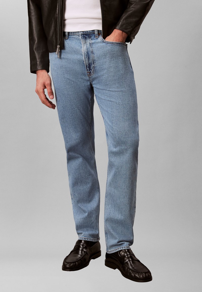 Άνδρας που φοράει ανοιχτό μπλε τζιν, μαύρα δερμάτινα μοκασίνια, λευκό πουκάμισο και σκούρο καφέ δερμάτινο μπουφάν, με το χέρι στην τσέπη, στέκεται πάνω σε γκρι φόντο.