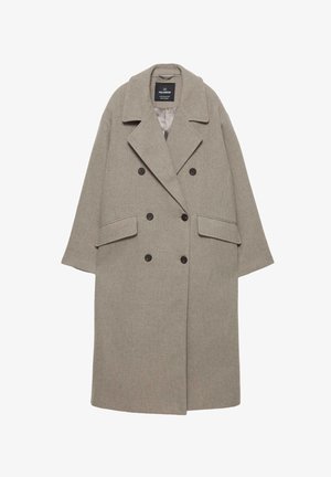 Osoba ubrana w długi beżowy płaszcz, niebieskie rozszerzane dżinsy i czarne buty, stojąca tyłem na jednolitym jasnym tle.