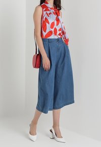 Bluzka bez rękawów w czerwony liściasty wzór na białych paskach, połączona z luźnymi niebieskimi dżinsowymi spodniami culottes. Kobieta trzyma małą czerwoną torebkę i ma na sobie białe szpilki.