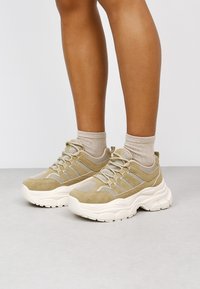 Baskets en mesh et en daim beige avec semelle en caoutchouc blanc, présentant des accents texturés et un design à lacets. Portées avec des chaussettes de couleur claire.