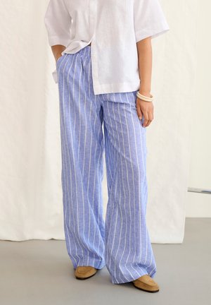 Losse blauwe broek met witte verticale strepen, gedragen met een wit shirt, beige schoenen en lichtgekleurde armbanden aan de pols.