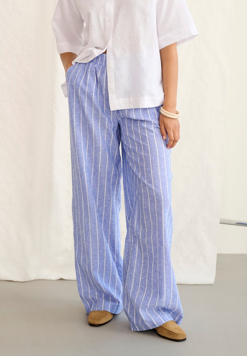 Voľné modré nohavice s bielymi vertikálnymi pruhmi, nosené s bielou košeľou, béžovými topánkami a svetlými náramkami na zápästí.