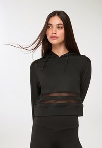 Giovane donna con lunghi capelli scuri che indossa un top nero con cappuccio e dettagli in pizzo, abbinato a leggings neri, su uno sfondo chiaro e semplice.