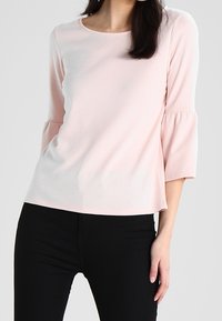 Světle růžový, měkký top s kulatým výstřihem a tříčtvrtečními rukávy s nařasenými detaily. V kombinaci s černými přiléhavými kalhotami.