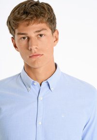 Lichtblauw button-up shirt met een kraag, gemaakt van stof met een gladde textuur. Heeft witte knopen en een klein geborduurd logo op de borst.