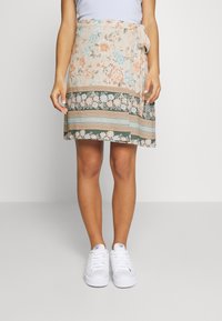 Saia envolvente floral em bege com flores azuis e pêssego. Apresenta uma faixa atada na cintura e uma bainha em camadas com padrão. Combinada com ténis brancos.