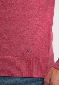 Pull en maille rouge présentant un ourlet et des poignets côtelés, avec un petit logo marron près du bas. Texture lisse et poids moyen.