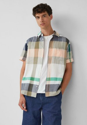 Junger Mann, der ein kurzärmeliges, mehrfarbig kariertes Hemd über einem weißen T-Shirt und blauen Jeans trägt, stehend vor einem schlichten hellen Hintergrund.