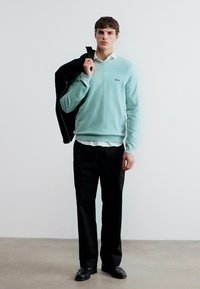 Jeune homme portant un pull bleu clair sur une chemise blanche, un pantalon noir et des chaussures noires, tenant une veste noire sur l'épaule.