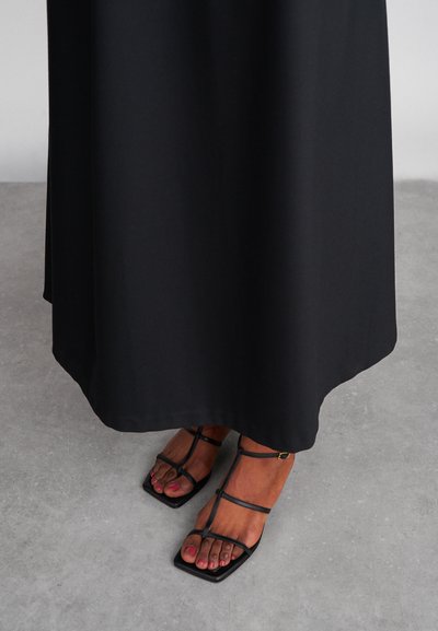 Vestido maxi preto com uma textura suave, combinado com sandálias pretas de tiras com biqueira quadrada e tiras finas, sobre um chão cinza.