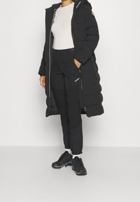 Musta pitkä untuvatakki vetoketjulla, hupulla ja monimutkaisella ompelulla, yhdistettynä mustiin housuihin ja tukeviin mustiin lenkkikenkiin.