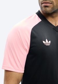 Mees, kes kannab musta spordisärki roosade varrukatega ja roosat Adidas trefoil-logot rinnal, vaadatuna veidi paremale.